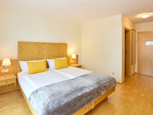 Hell eingerichtetes Doppelzimmer mit Holzmöbeln gelben Kissen und grauer Bettdecke auf Holzboden