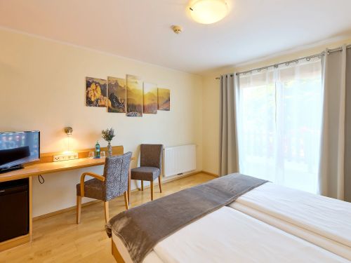 Helles hotelzimmer mit zwei betten einem schreibtisch zwei sesseln und einem fenster mit gardinen und bergbild an der wand