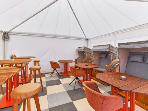 Sitzbereich in einem weißen zelt mit holztischen hohen hockern orangenen stuhlen und geflochtenen strandkorbstühlen
