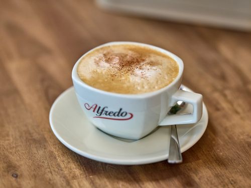 Weisse Tasse mit Cappuccino und Milchschaum auf weissem Teller mit Löffel auf Holztisch
