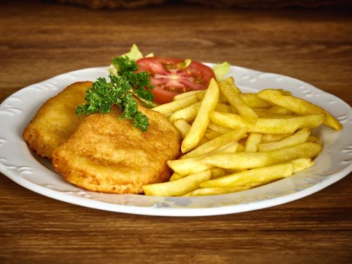 Schnitzel mit pommes und tomatenscheibe garniert mit petersilie auf weissem teller vor holzhintergrund