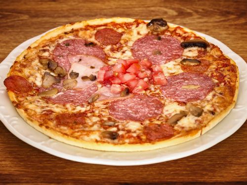 Pizza mit Salami Schinken Pilzen und frischen Tomaten auf weißem Teller vor Holzuntergrund