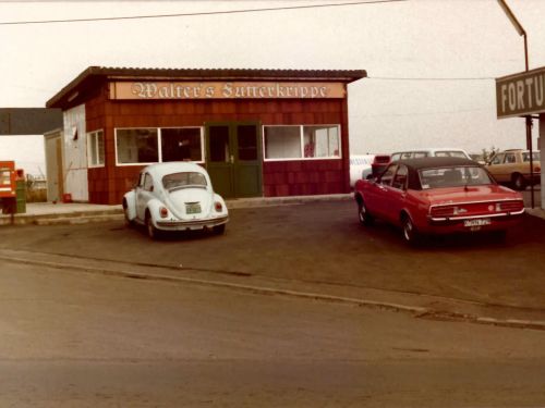 Parkplatz vor schnellem Imbiss mit zwei alten Autos und Werbeschildern in leicht trüber Umgebung