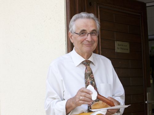 Älterer Mann in weißem Hemd mit gemusterter Krawatte hält Wurst im Brötchen vor Holztür mit Schild Zutritt nur für Personal