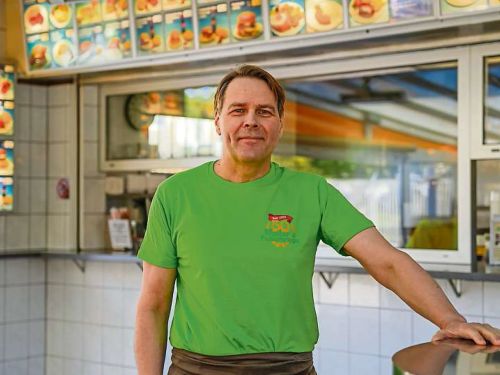 Mann in grunem tshirt steht in imbissbude vor bestelltheke mit speisekarten im hintergrund